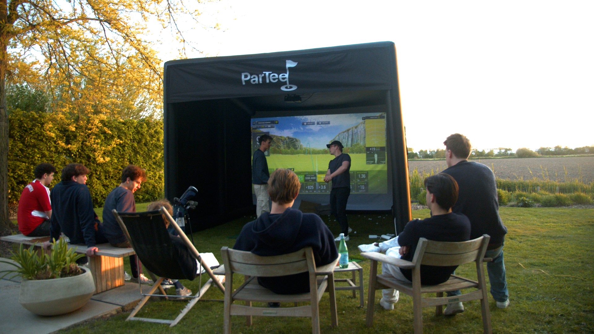 Friends gathered around the ParTee mobile golf simulator in a garden at sunset